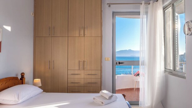 A bedroom with a bed, a wooden wardrobe, a balcony with a view of the sea, and a window with curtains. - Albania Dream Saranda Apartments (Sarandë)