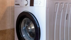 A front-loading washing machine in a bathroom with tiled walls and a white towel hanging on the wall. - Albania Dream Saranda Apartments (Sarandë)