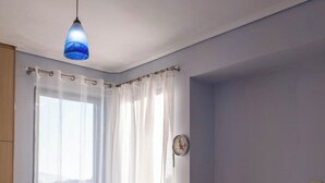 A bedroom with two beds, a bedside table with a lamp, a window with sheer curtains, and a clock on the wall. - Albania Dream Saranda Apartments (Sarandë)