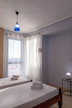 A bedroom with two beds, a bedside table with a lamp, a window with sheer curtains, and a clock on the wall. - Albania Dream Saranda Apartments (Sarandë)