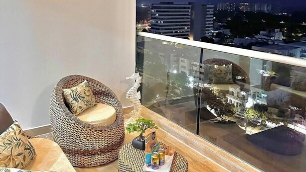 Un balcón con mobiliario de mimbre, una pequeña mesa con bebidas y vistas a la ciudad de noche.