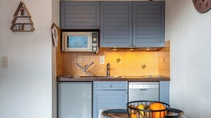 Une cuisine compacte avec des meubles gris, un four à micro-ondes, un évier et une petite table sur laquelle se trouve un panier de fruits.