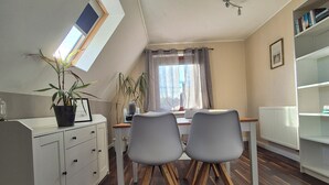 Een eetgedeelte met een tafel, twee stoelen, een potplant, een raam met gordijnen en een boekenkast.