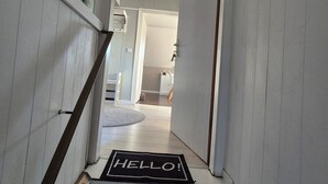 Een gang met een 'HELLO!' mat aan het einde, een trap met een leuning, en een deur die naar een andere kamer leidt.