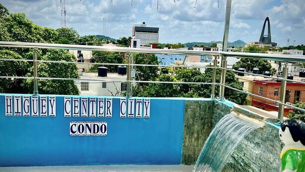 Une piscine agrémentée d’une cascade, une enseigne indiquant « HIGUEY CENTER CITY CONDO », et une vue sur le paysage urbain environnant.