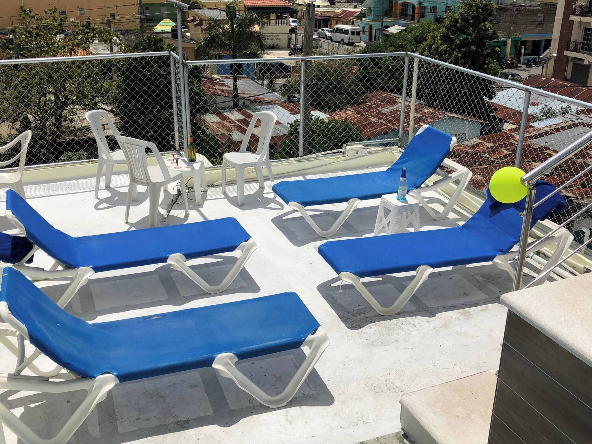 Terraza en la azotea con sillones azules y una pequeña mesa, rodeada por una cerca de malla metálica.