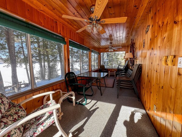 Interior de cabana com paredes de madeira, ventilador no teto, mesa de jantar com cadeiras, sofá e janelas amplas com vista para a neve.