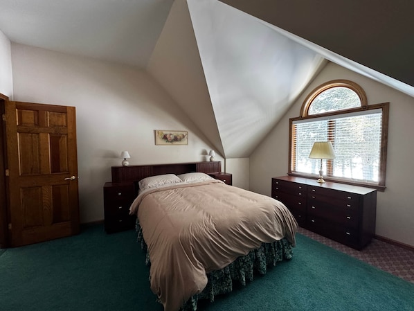 Ein Schlafzimmer mit schrägen Wänden, einem Bett mit Blumenmuster, einer Kommode mit Lampe und einem Fenster mit Jalousien.