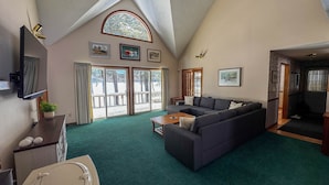 A high-ceilinged living room with a large sofa, a flat-screen TV, and a view through the glass doors to the outside. - Lakefront Peninsula Retreat w/Private Dock, on Cobbosseecontee Lake, 4 BR (West Gardiner)