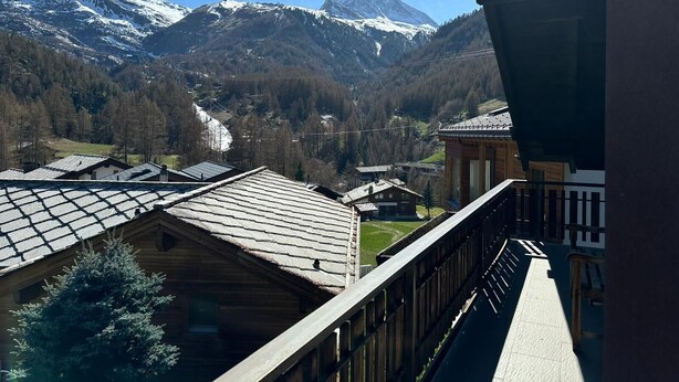 Une vue sur les montagnes, avec des sommets enneigés, un bâtiment en bois doté d’un toit en pente et un balcon donnant sur le paysage.