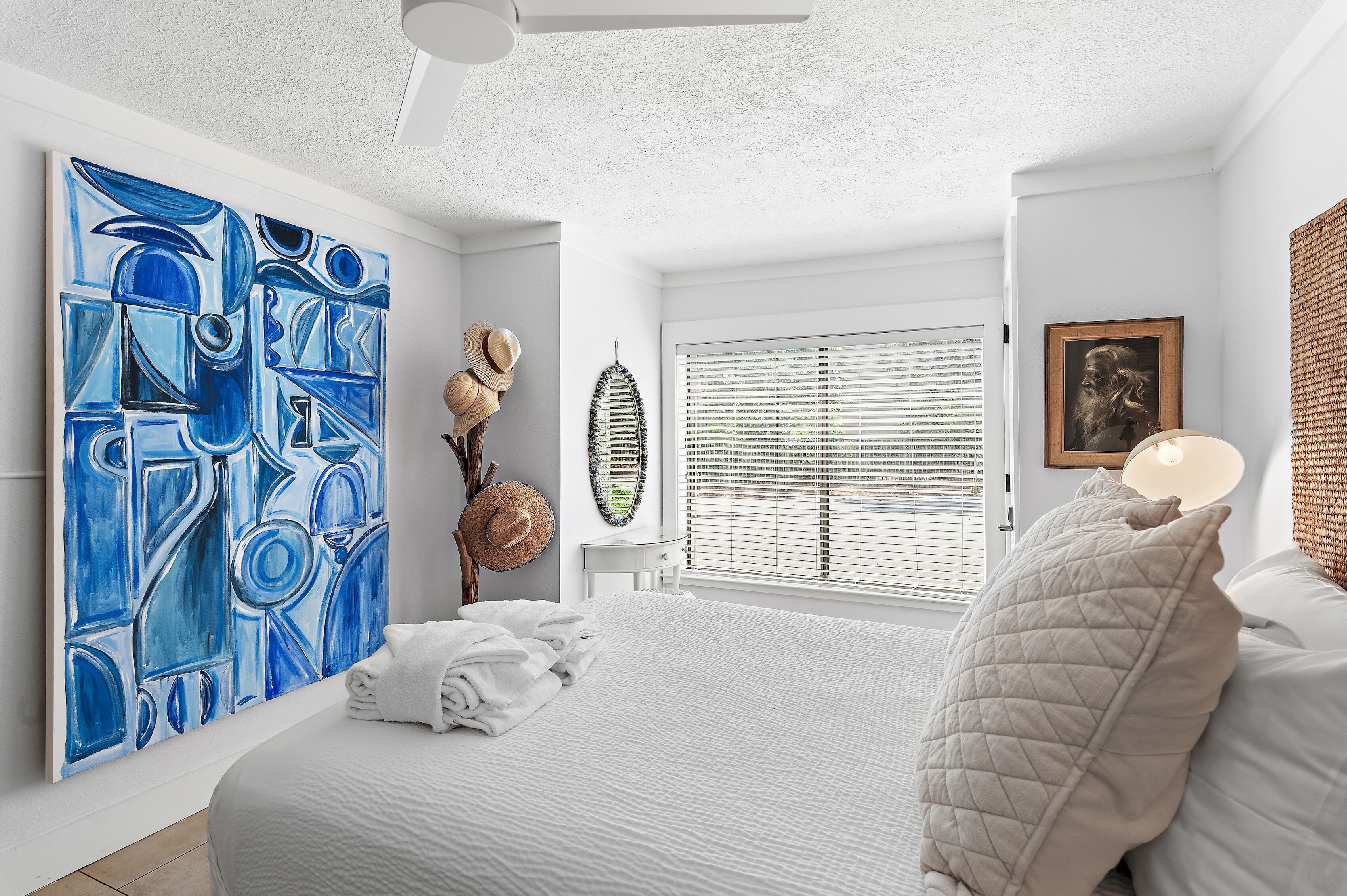 Un dormitorio con un cuadro abstracto grande, una cama con ropa blanca, una ventana con persianas y un ventilador de techo.