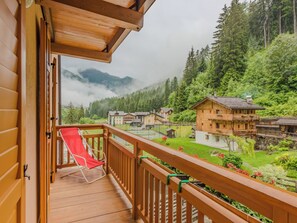 Un balcon en bois avec une chaise rouge, offrant une vue sur un paysage montagneux parsemé de maisons traditionnelles en bois et une vue dégagée sur la vallée en contrebas.