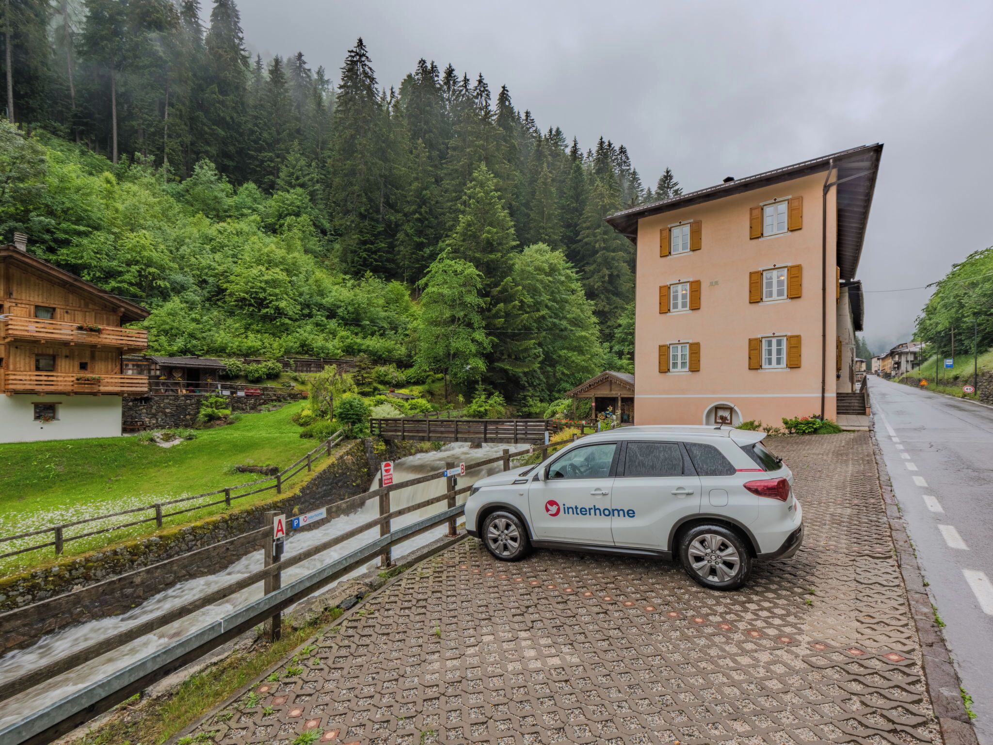 A white car parked on a cobblestone path, a building with a sloped roof, and a forested hillside in the background.