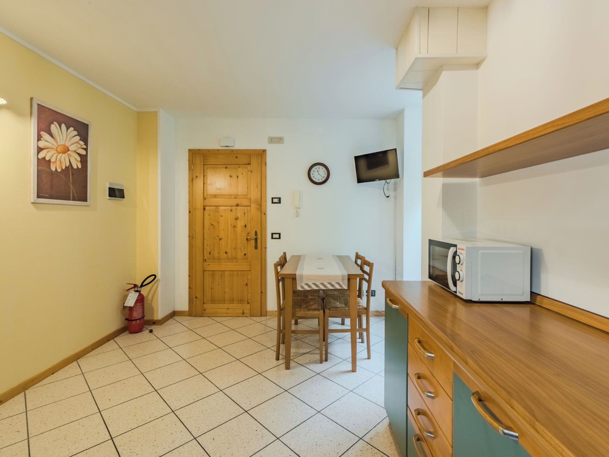 Une petite cuisine avec une table en bois, des chaises, un four à micro-ondes, une horloge et un extincteur.