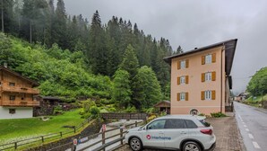 Ein weißes Auto parkt auf einem Kopfsteinpflasterweg, ein Gebäude mit geneigtem Dach und ein bewaldeter Hang im Hintergrund.