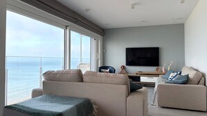 A modern living room with a large window overlooking the sea, a flat-screen TV mounted on the wall, and a sofa with a throw blanket. - Apartment With sea View in Middelkerke for 6 (Middelkerke)