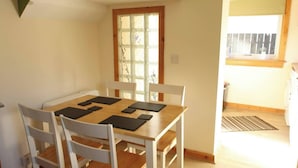 A dining area with a wooden table and white chairs, a window with a view, and a kitchen visible in the background. - House on the shores of Loch Shiel in Historic Glenfinnan, Monument and Viaduct (Glenfinnan)