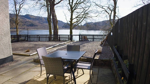 An outdoor patio with a table and chairs, a wooden fence, and a view of a lake and hills. - House on the shores of Loch Shiel in Historic Glenfinnan, Monument and Viaduct (Glenfinnan)