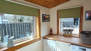 A laundry area with a washing machine, dryer, and a window with a view of a wooden fence and trees. - House on the shores of Loch Shiel in Historic Glenfinnan, Monument and Viaduct (Glenfinnan)