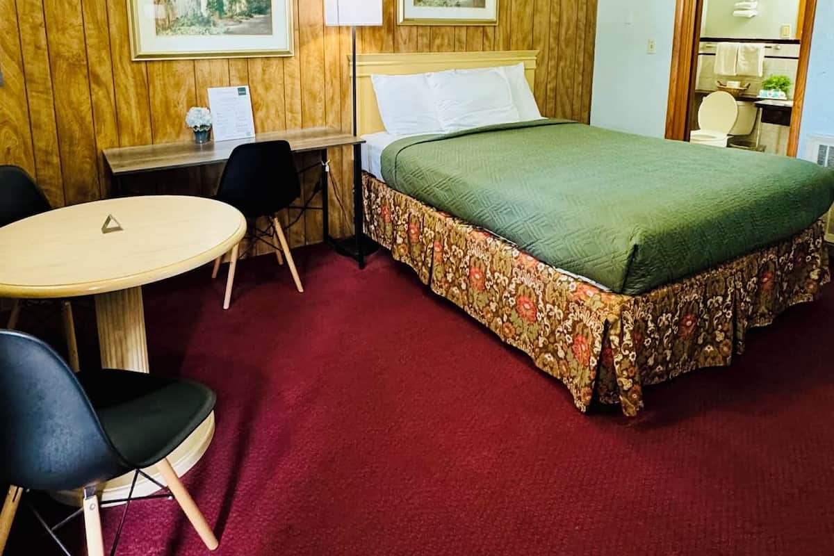 A hotel room with a wooden paneled wall, a bed with a floral bedspread, a desk with a chair, a round table, and a mirror.