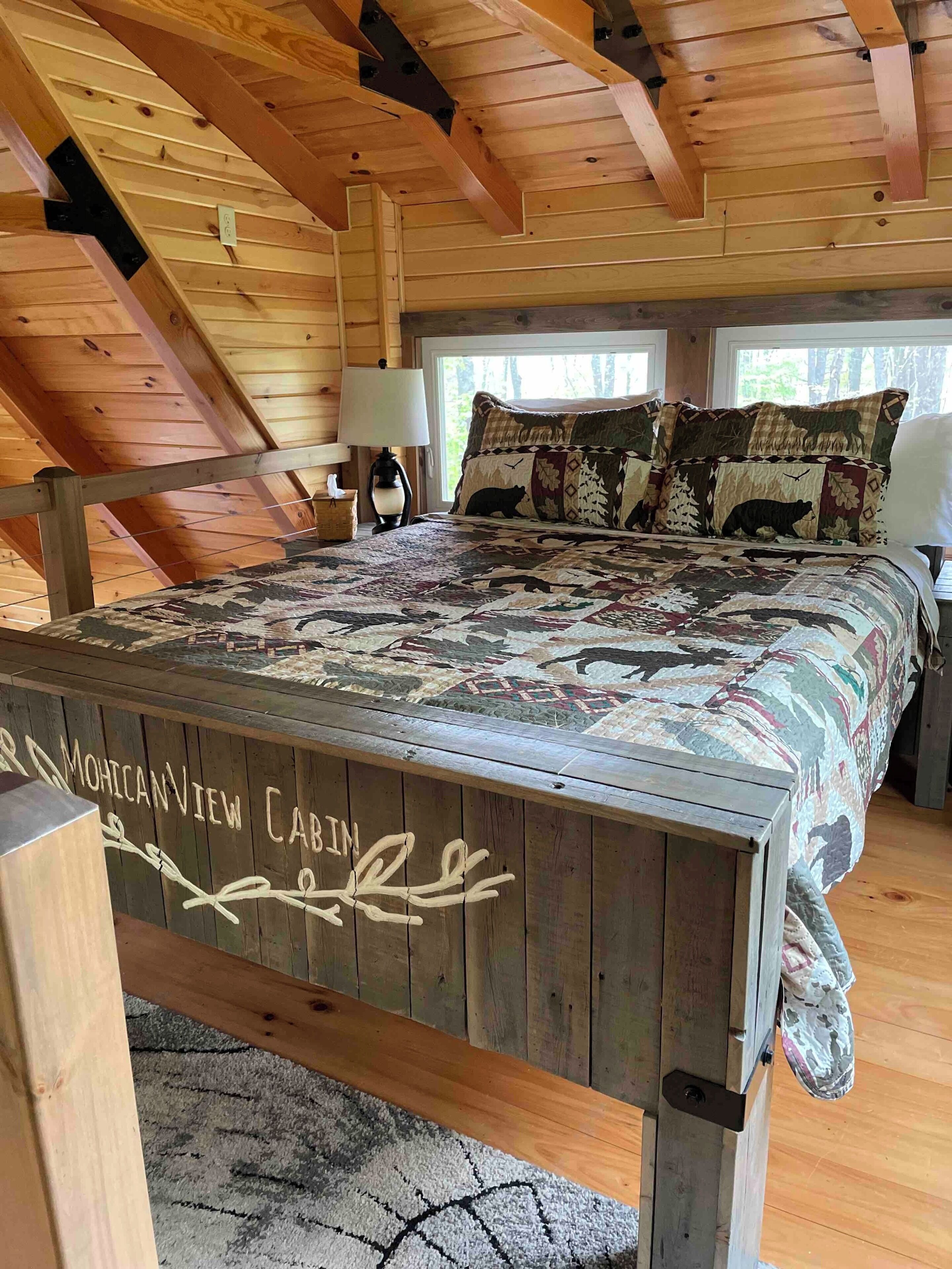 Ein Interieur einer Holzhütte mit einem hölzernen Bett, einem Nachttisch, einer Lampe und Blick ins Freie durch ein Fenster.