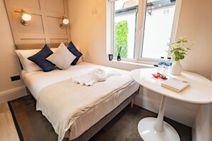 Een net opgemaakt bed met kussens, een nachtkastje met een boek en een vaas, en een raam met uitzicht op groen.