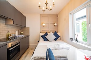 A modern hotel room with a kitchenette, a bed with white and blue bedding, a chandelier, and a window with a view.