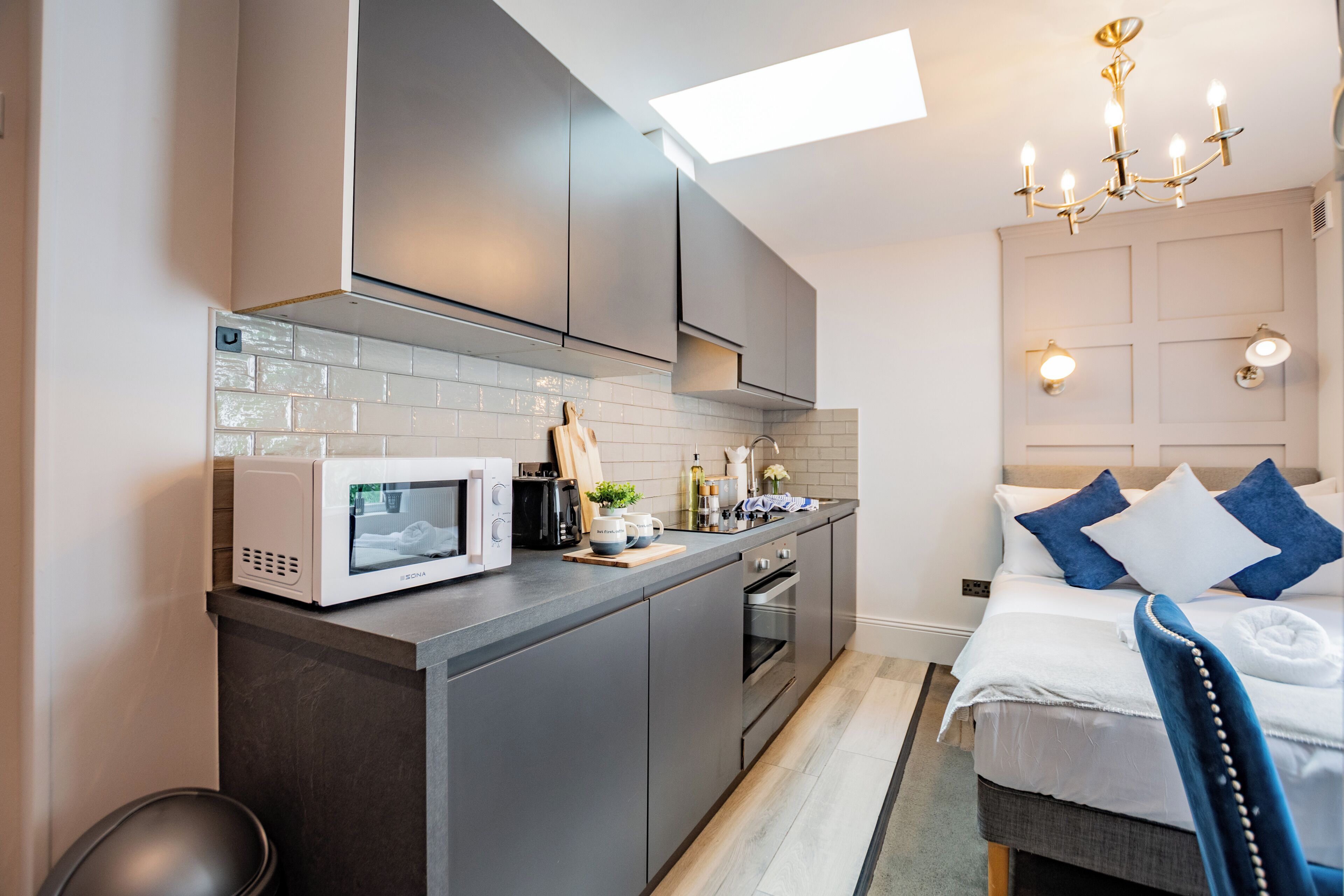 A modern kitchen with a microwave, toaster, and oven, a small dining area, and a bedroom with a bed and nightstand.