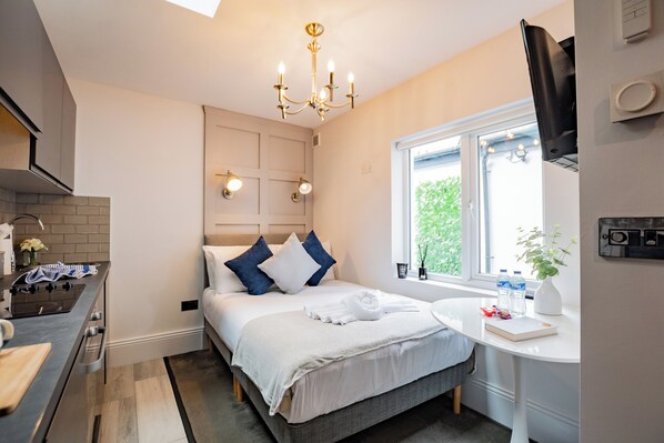 Een moderne hotelkamer met kitchenette, een bed met wit en blauw beddengoed, een nachtkastje met een boek en een vaas, en een raam met uitzicht op groen.