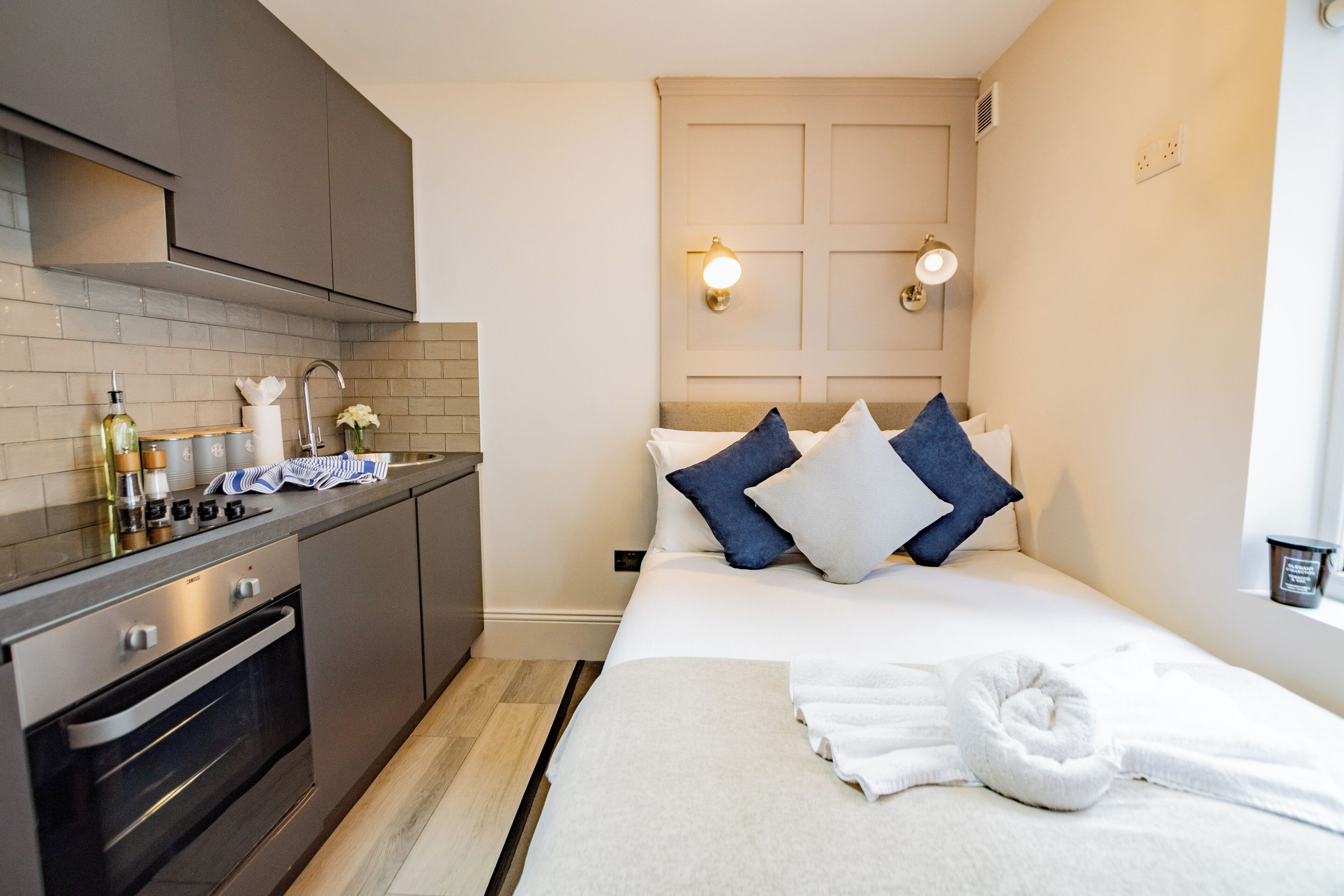 Habitación de hotel compacta con kitchenette, una cama con almohadas, una toalla enrollada y una lámpara fijada a la pared.