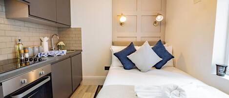 Habitación de hotel compacta con kitchenette, una cama con almohadas, una toalla enrollada y una lámpara fijada a la pared.