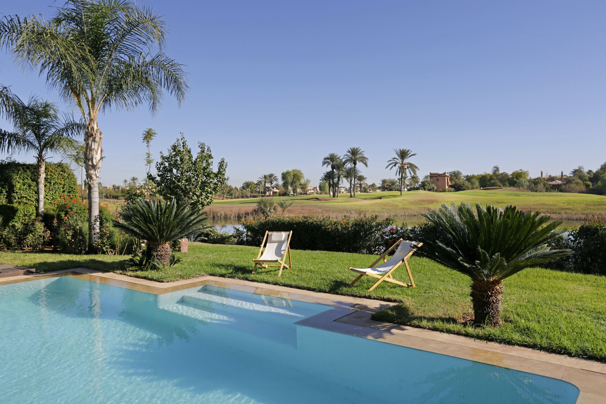 Ein Swimmingpool mit zwei Liegestühlen, Palmen und Blick auf einen Golfplatz.