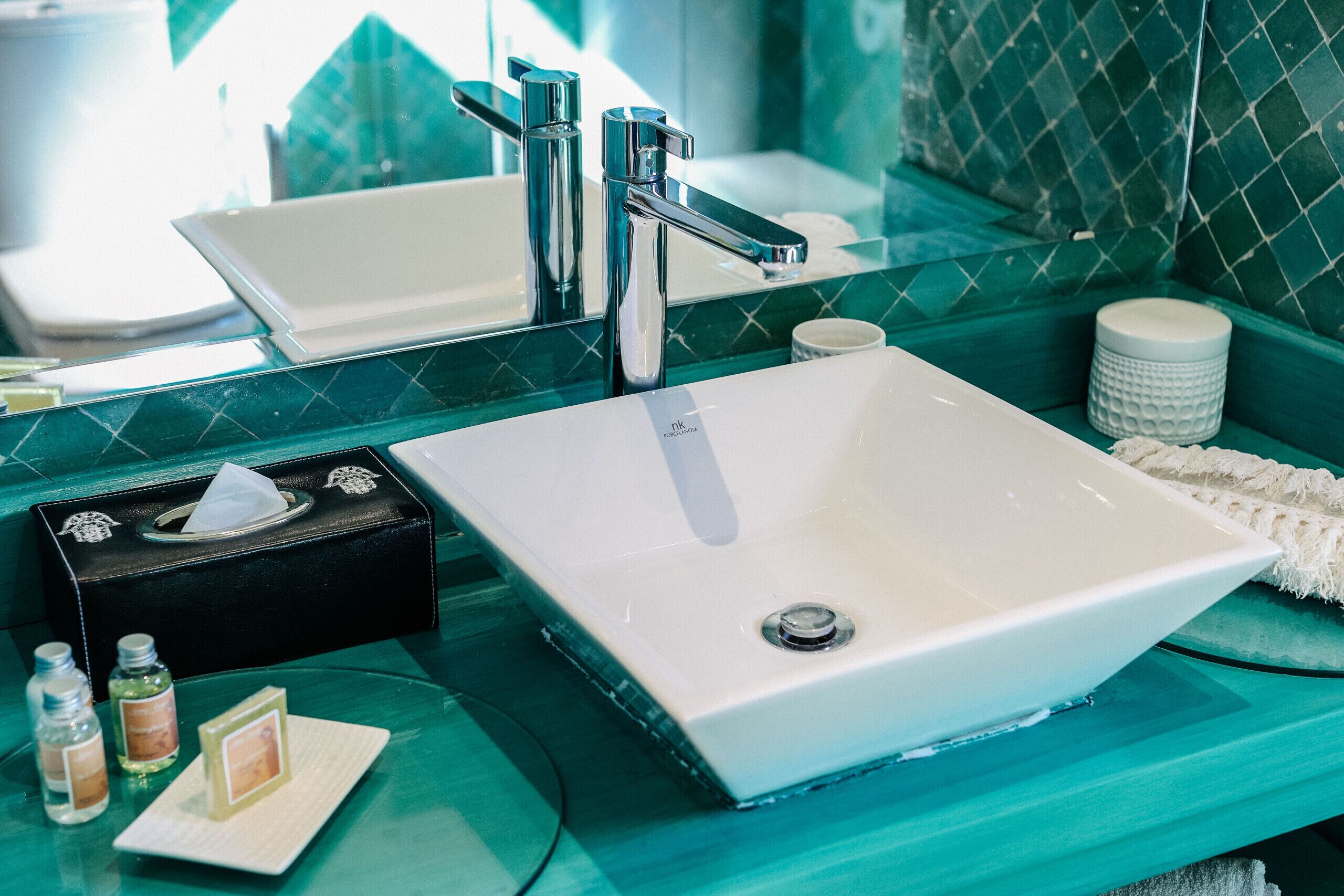 Un baño con lavamanos blanco, grifo cromado y un espejo que refleja una pared con azulejos verdes.