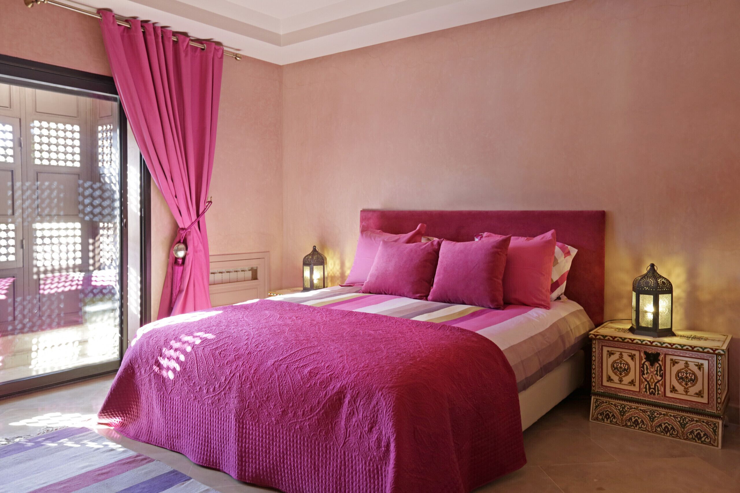 A bedroom with a bed, pink curtains, a nightstand with a lantern, and a view through a glass door.