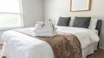 Una cama bien hecha con sábanas blancas y una manta marrón, una pila de toallas dobladas y una cita enmarcada en la pared.