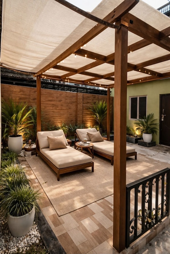 A covered outdoor seating area with cushioned benches and tables, surrounded by potted plants and a wooden fence.