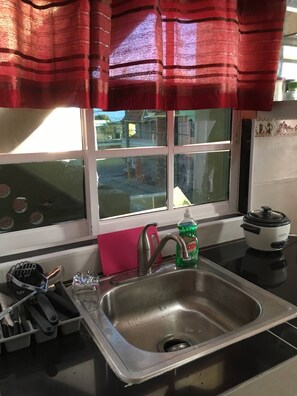 A kitchen with a stainless steel sink, a window with a view of the street, and a red curtain above the sink. - Higuey Center City, Leslie's Villa (Higuey)