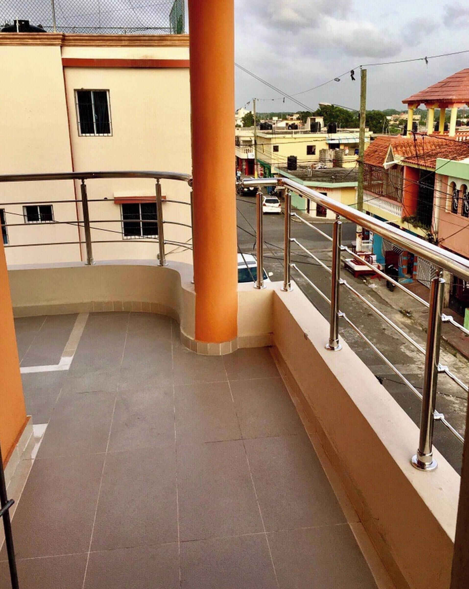 A balcony with a metal railing, a tiled floor, and a view of a street with buildings and parked cars.