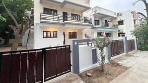 A modern two-story residential building with a balcony, a gated entrance, and a small garden area. - Nisarg Homestay with Kitchen (Bhuj)