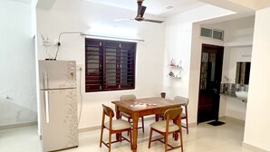 A dining area with a table and chairs, a refrigerator, a window with blinds, and a ceiling fan. - Nisarg Homestay with Kitchen (Bhuj)
