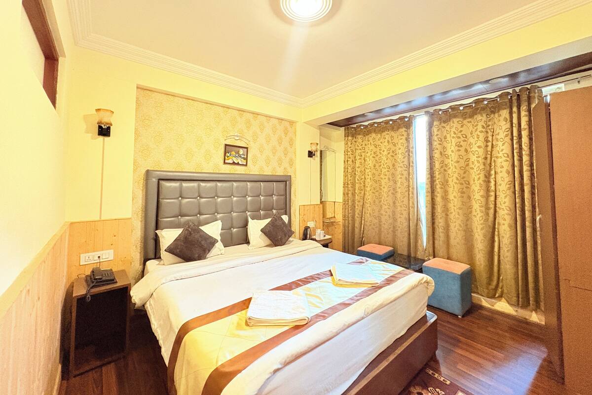 Habitación de hotel con una cama grande, mesitas de noche, una pequeña mesa, un espejo y una ventana con cortinas.