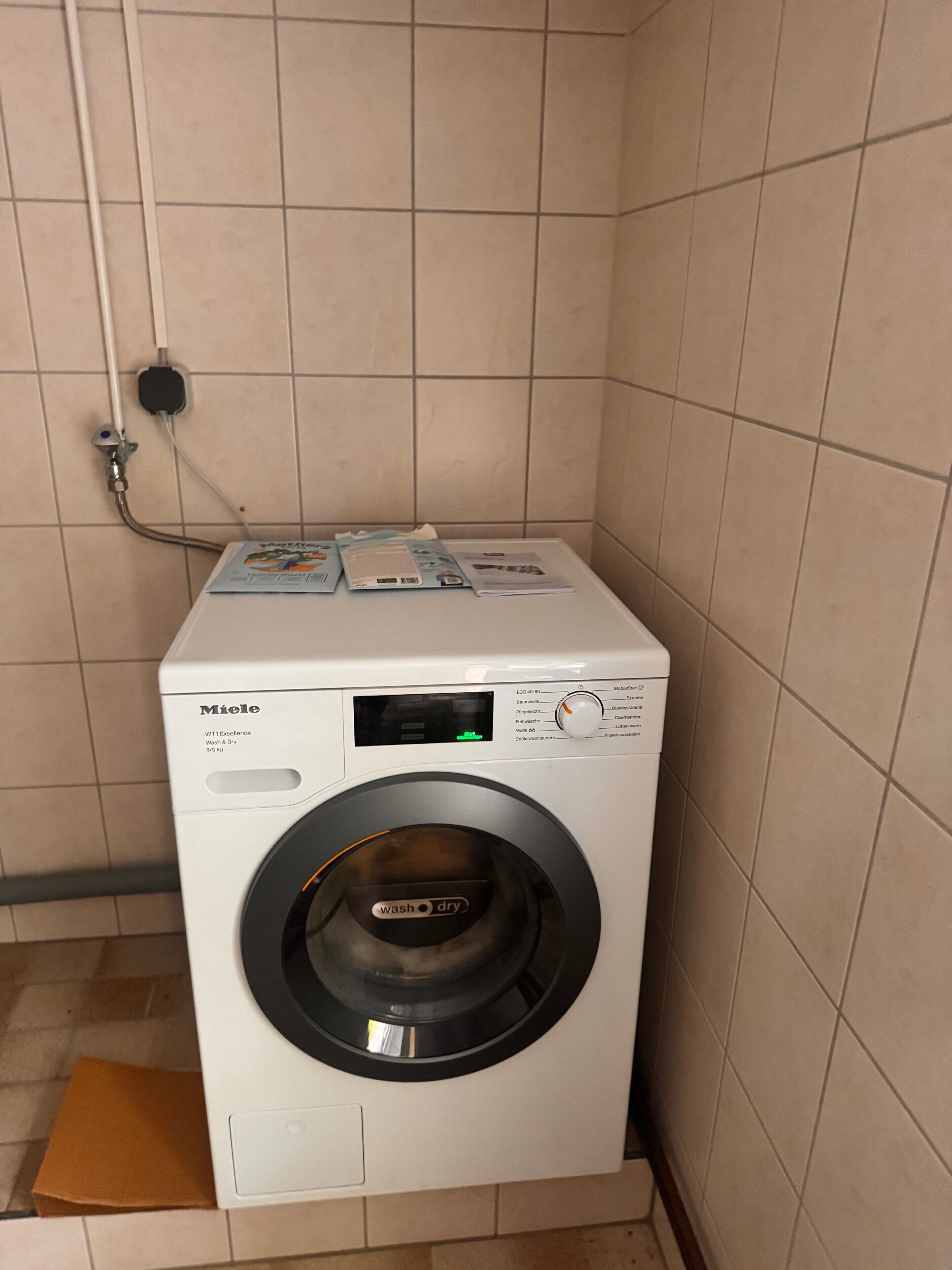 A white Miele washing machine in a tiled room with a control panel and a power outlet on the wall.