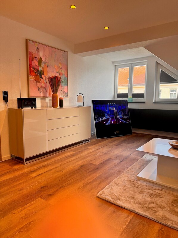 Una sala de estar moderna con piso de madera, televisor de pantalla plana, mueble blanco, un cuadro y un jarrón con flores secas.