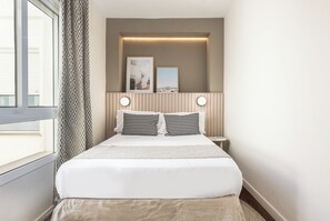 Ein ordentlich eingerichtetes Hotelzimmer mit einem großen Bett, einem Nachttisch, einem Beistelltisch und einem Fenster mit Vorhängen.