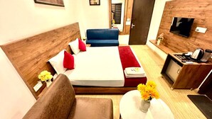 Ett hotellrum med en säng, en soffa, ett litet bord med en vas med blommor, en tv och ett skrivbord med en kaffebryggare.
