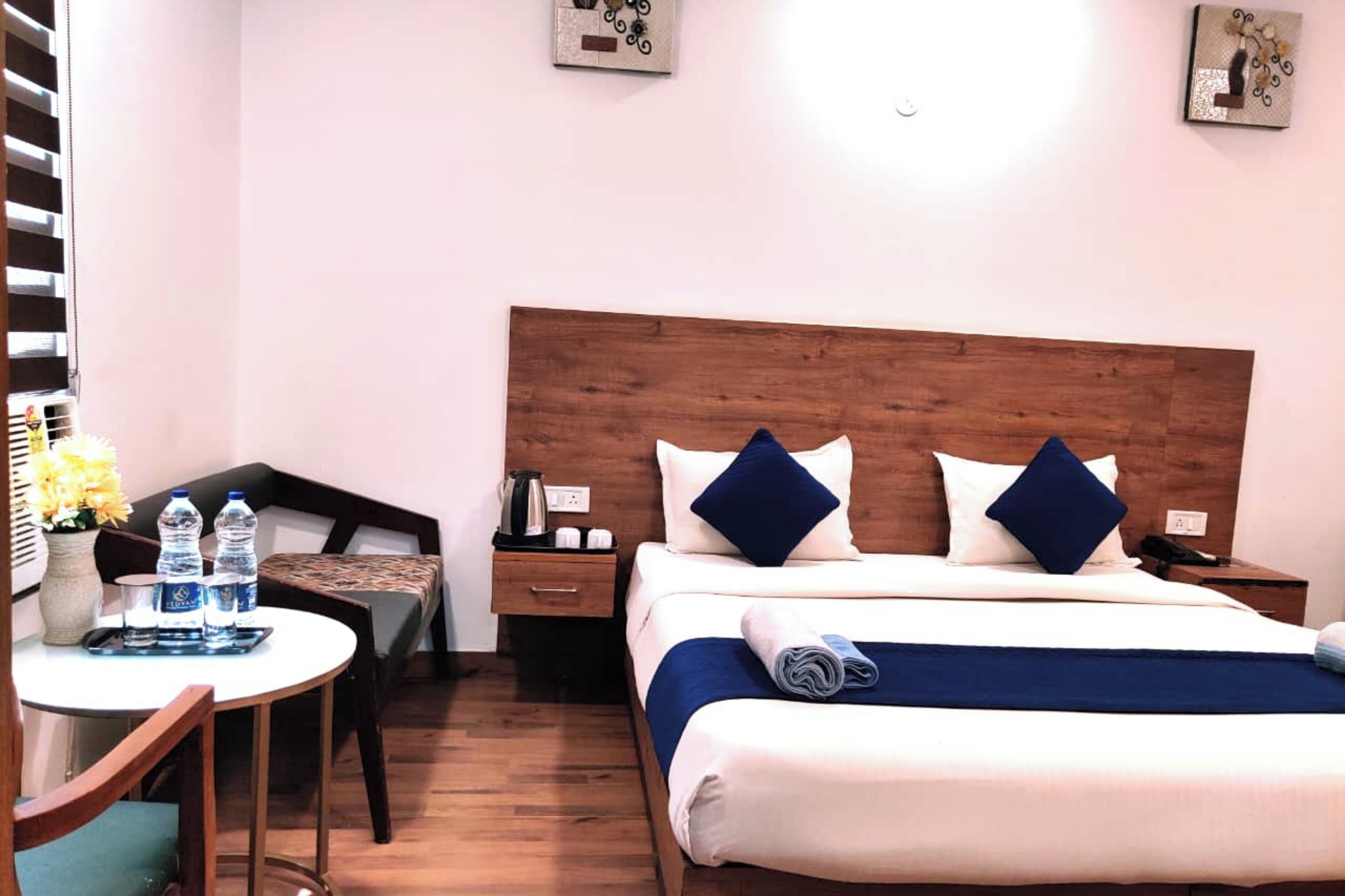 Una camera d'albergo con una testiera in legno, un letto con biancheria azzurra e bianca, un tavolino con bottiglie d'acqua, una sedia e un comodino con un telefono e una lampada.