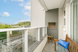 A balcony with a glass railing, a wooden chair, a small table with a bottle, and a blue pillow with 'blue check-in' text. - Modern | Fast Wi-Fi | On the Gastronomic Route (Florianópolis)