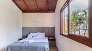 Ein Schlafzimmer mit Bett, Nachttisch, Fenster und Blick auf Palmen sowie einer Holzbalkendecke.