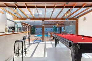 A pool table with a red felt surface, a bar area with stools, and a pool table in the background. - Beachfront house: pool, BBQ, jacuzzi & more (Florianópolis)