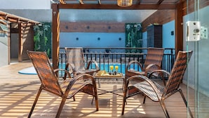 Eine Holzterrasse mit Korbstühlen und einem Tisch, im Hintergrund ein Pool und eine Glasbrüstung.